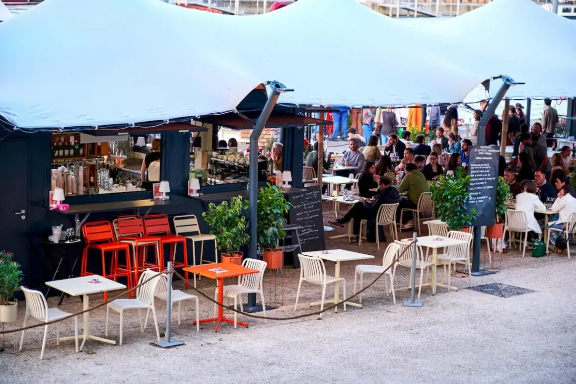 Bar animé sur les quais parisiens, avec des tables et des chaises en terrasse sous une grande tente blanche.