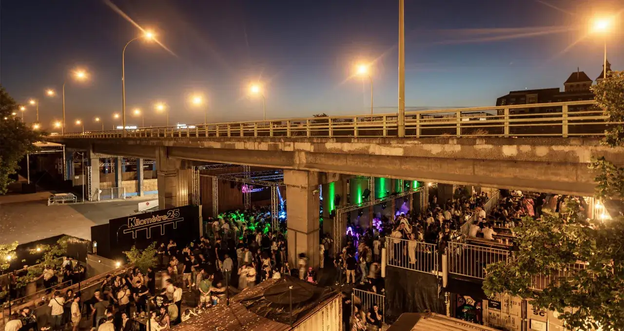 Soir&eacute;e anim&eacute;e sous un pont, avec une foule dansant au son de la musique. Le panneau 