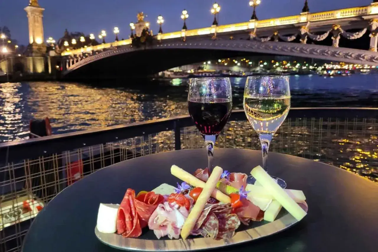 Un plateau de charcuterie et fromages avec deux verres de vin, sur fond de Seine illumin&eacute;e et du Pont Alexandre III. Une parfaite Riviera fuga parisienne.