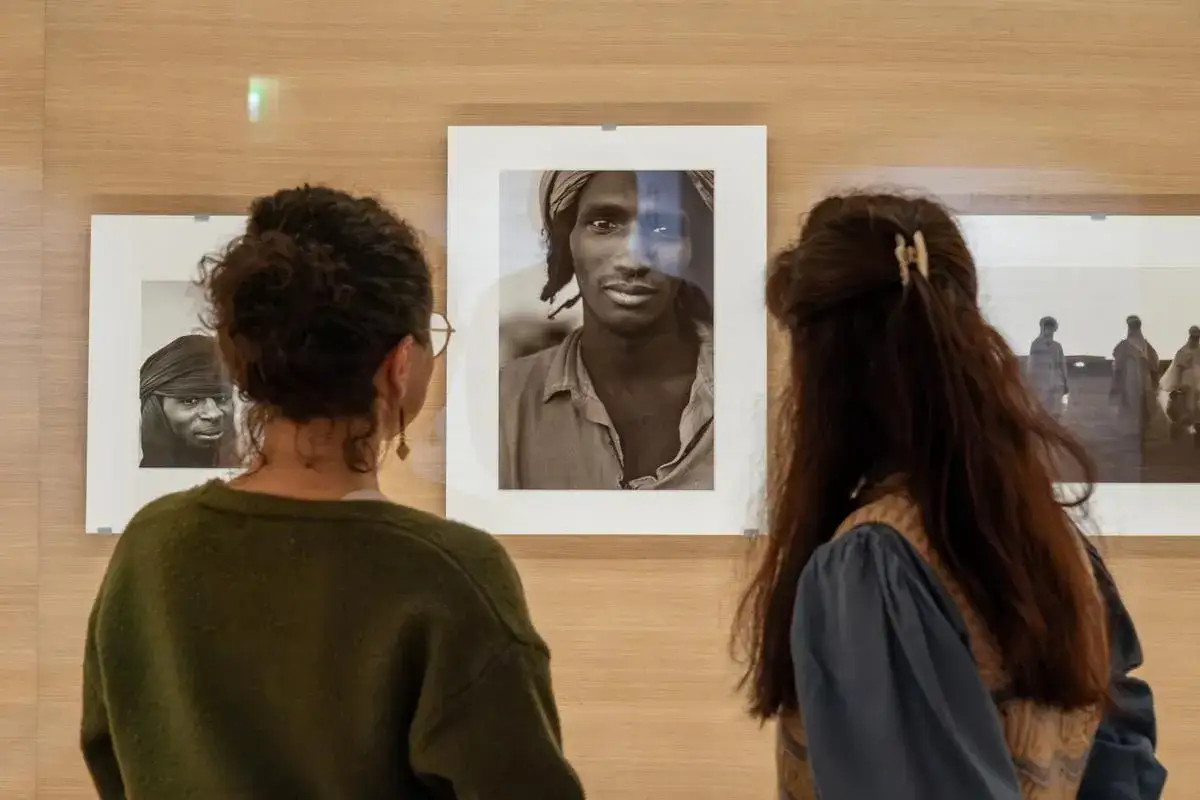 Deux femmes admirent des portraits saisissants lors d'une expo Paris en ce moment. L'une regarde un homme au turban, l'autre une sc&egrave;ne de figures lointaines.
