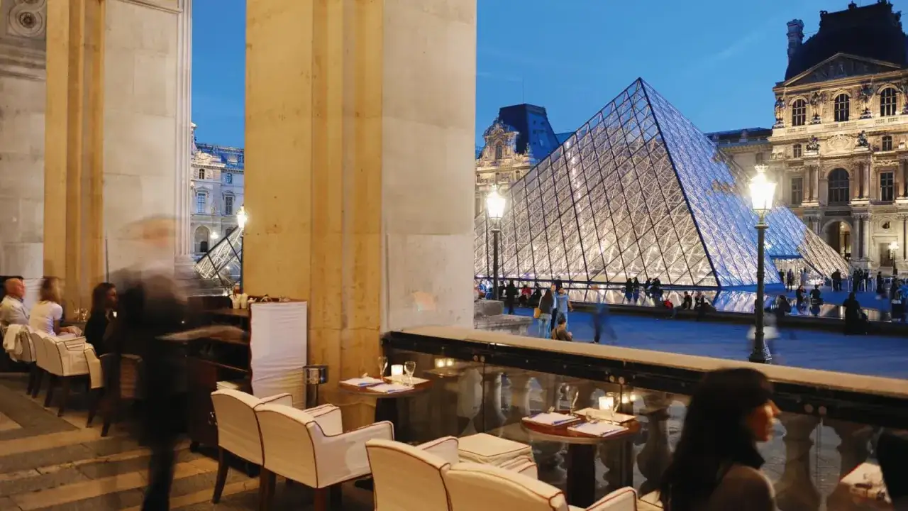 Terrasse du Café Marly, vue sur la pyramide du Louvre illuminée. Ambiance parisienne en soirée.