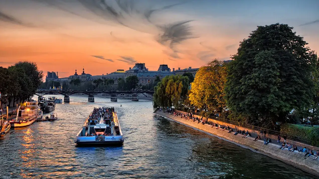 Coucher de soleil sur la Seine, un bateau plein de touristes navigue pr&egrave;s d'un bar sur les quais parisiens. Des gens profitent de la douceur du soir.