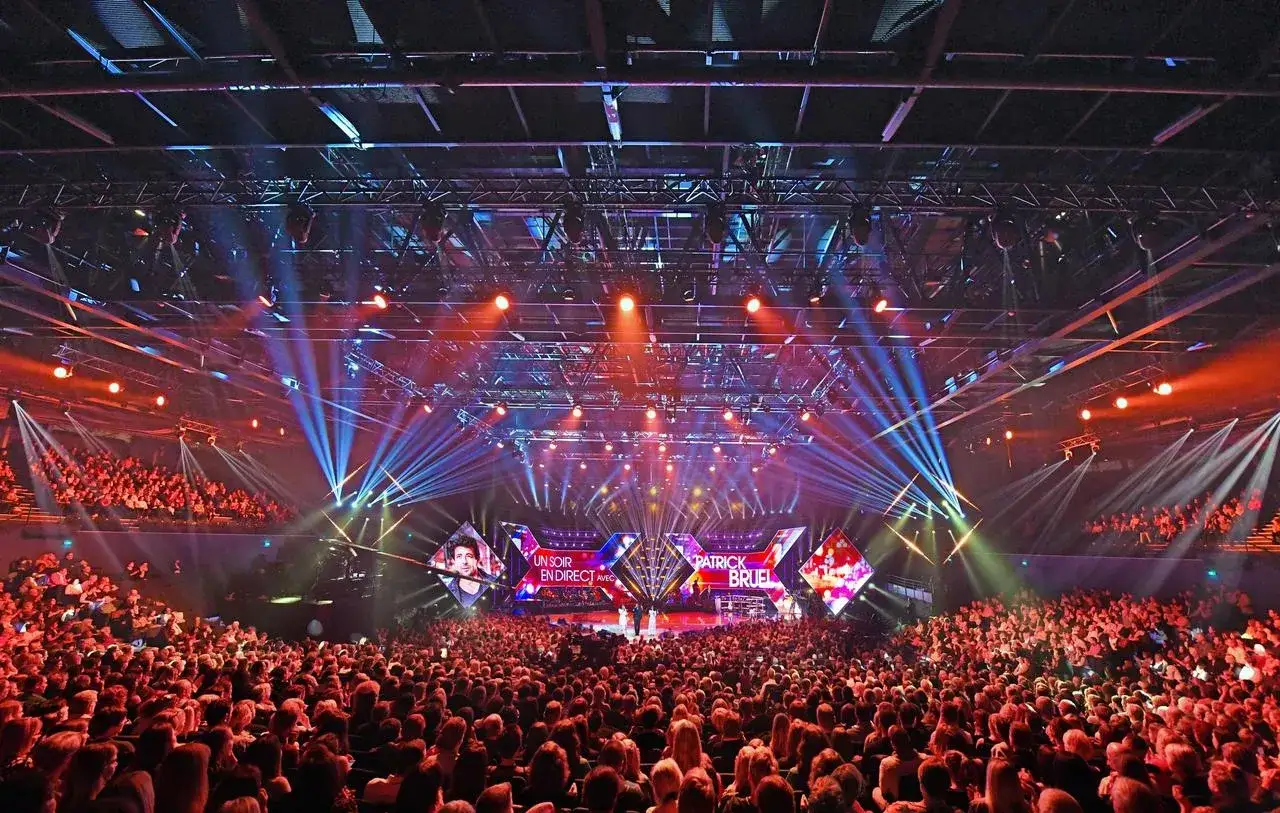 Sc&egrave;ne de concert spectaculaire avec Patrick Bruel, entour&eacute; de lumi&egrave;res vives et d'une foule immense, rappelant l'ambiance d'un grand spectacle au D&ocirc;me de Paris.