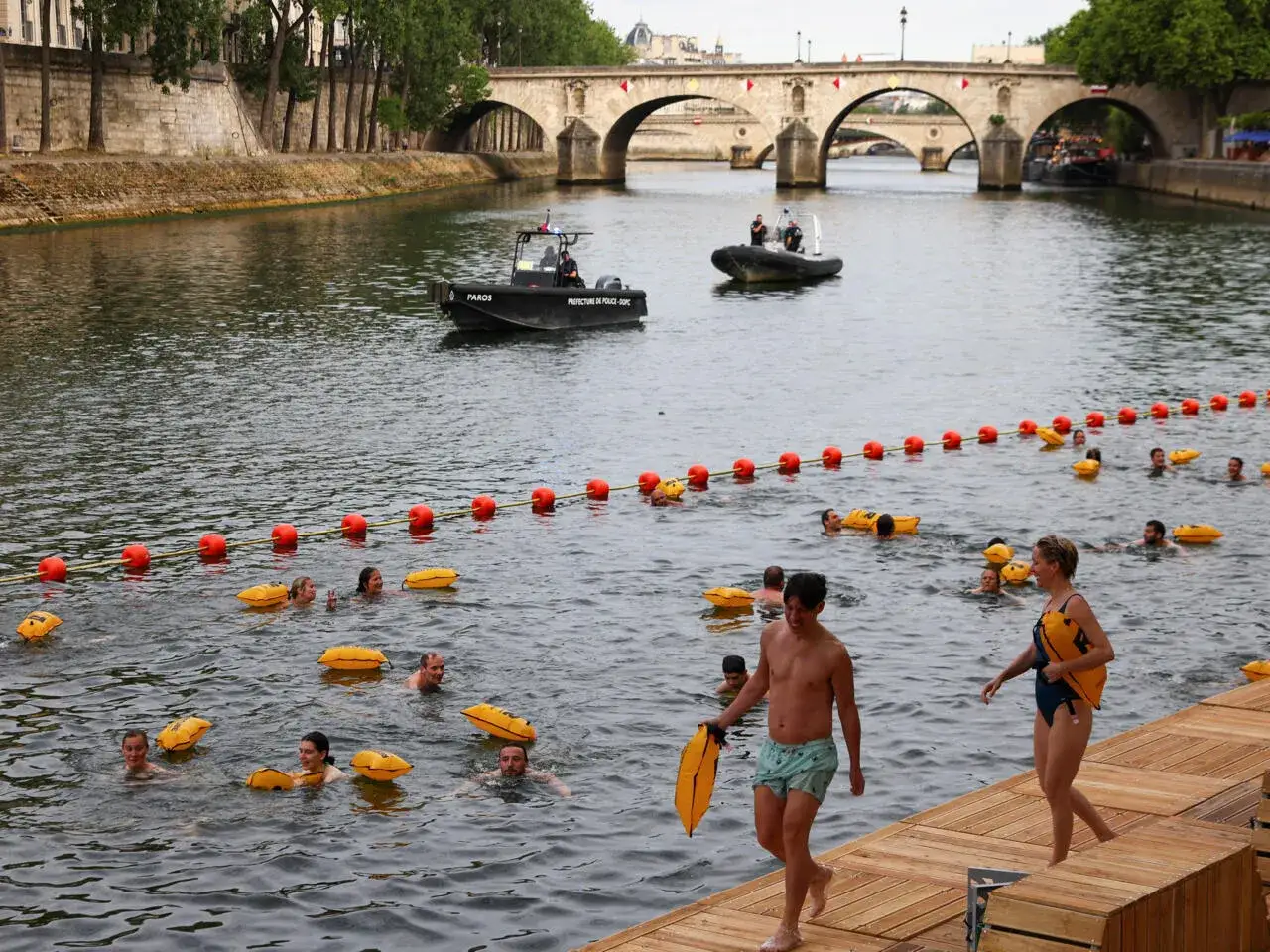 Des baigneurs profitent de la baignade dans la Seine, sous le regard de bateaux de police. Des bou&eacute;es orange d&eacute;limitent la zone.