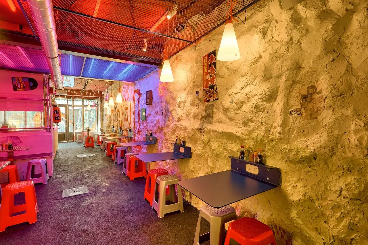 Ambiance néon et rock dans ce restaurant Noori Paris. Tables pliantes, tabourets colorés et murs texturés créent un décor unique.
