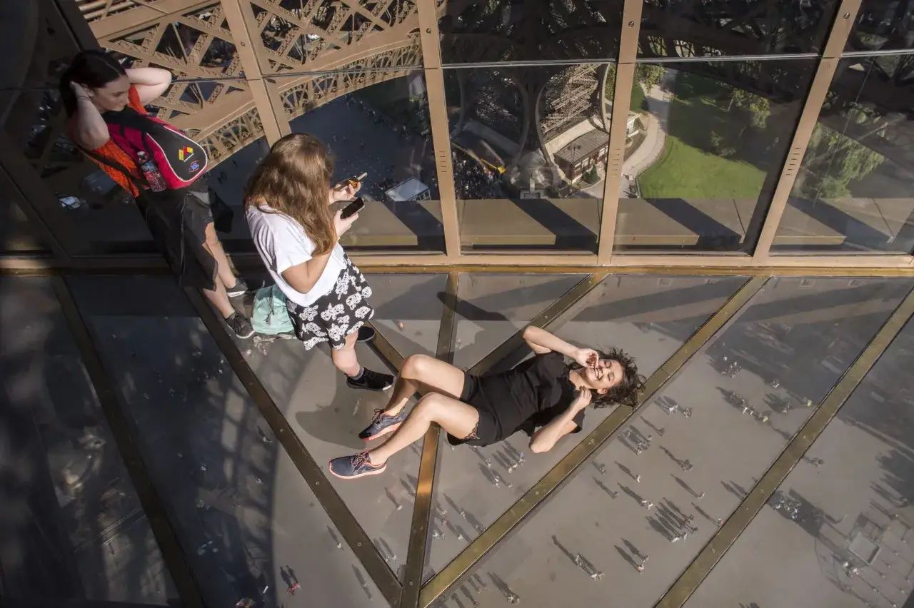 Vue plongeante sur le sol vitr&eacute; de l'ascenseur de la Tour Eiffel. Une jeune femme se repose, tandis que d'autres admirent le paysage parisien.