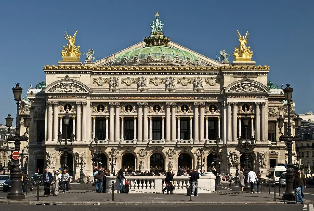 La place de l'Op&eacute;ra &agrave; Paris, avec son imposant b&acirc;timent orn&eacute; de statues dor&eacute;es et d'une coupole verte, sous un ciel bleu &eacute;clatant.