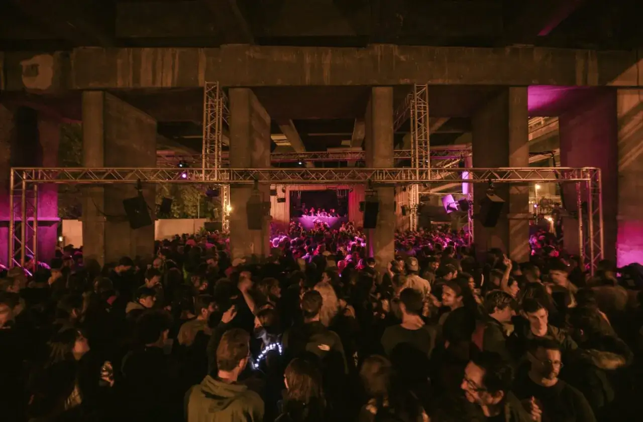Foule dansante sous un pont, ambiance &eacute;lectrique pour le virage paris. Lumi&egrave;res roses et sc&egrave;ne au loin.