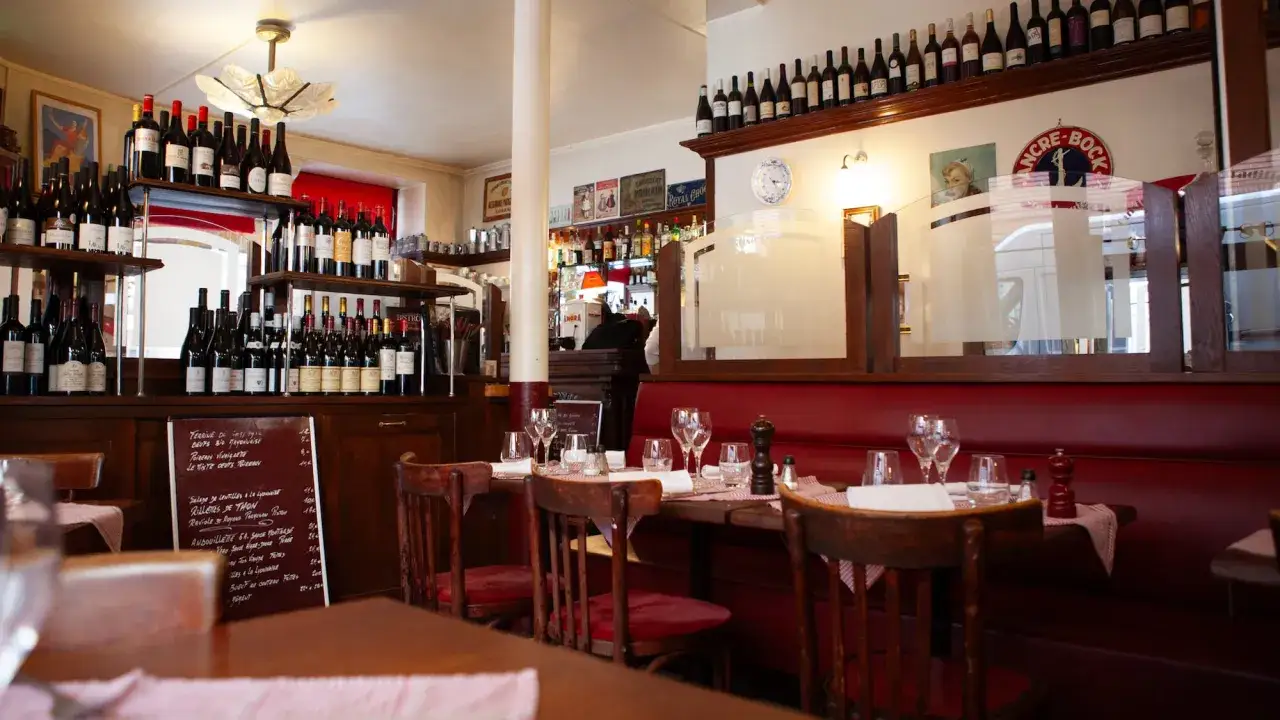 Int&eacute;rieur d'un bistrot parisien avec des &eacute;tag&egrave;res remplies de bouteilles de vin, tables dress&eacute;es, banquettes rouges. L'ambiance &eacute;voque le plus vieux restaurant de Paris.