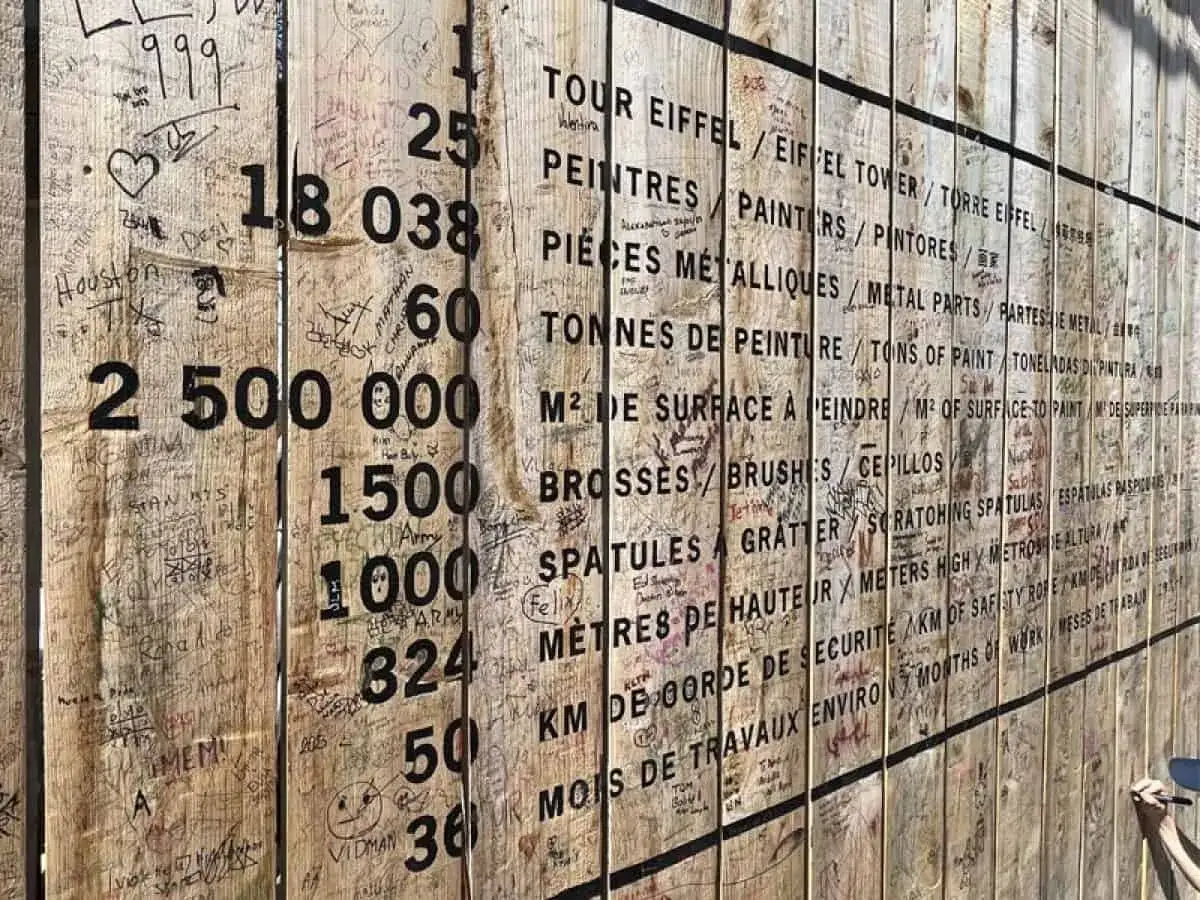 Panneau de chantier avec des chiffres et des mots sur la peinture de la Tour Eiffel. Des graffitis et des signatures recouvrent le bois.