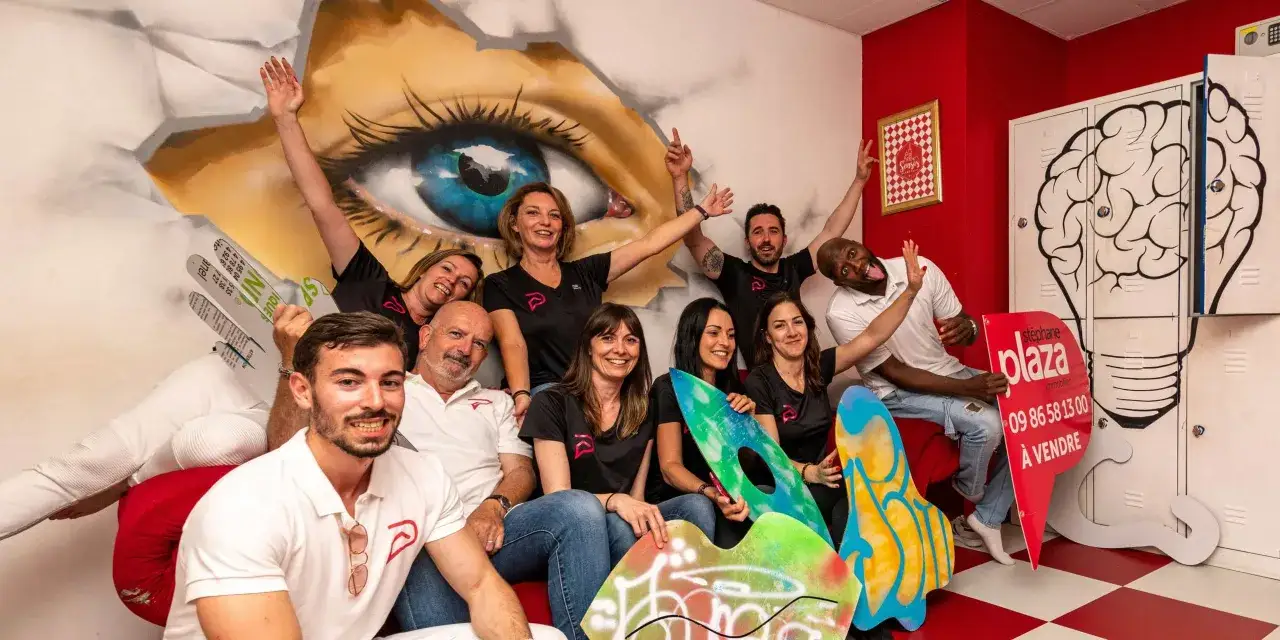 Une &eacute;quipe souriante pose devant un mur d&eacute;cor&eacute; d'un &oelig;il g&eacute;ant. Ambiance sensas Paris, avec des &eacute;l&eacute;ments graphiques color&eacute;s et un panneau immobilier.