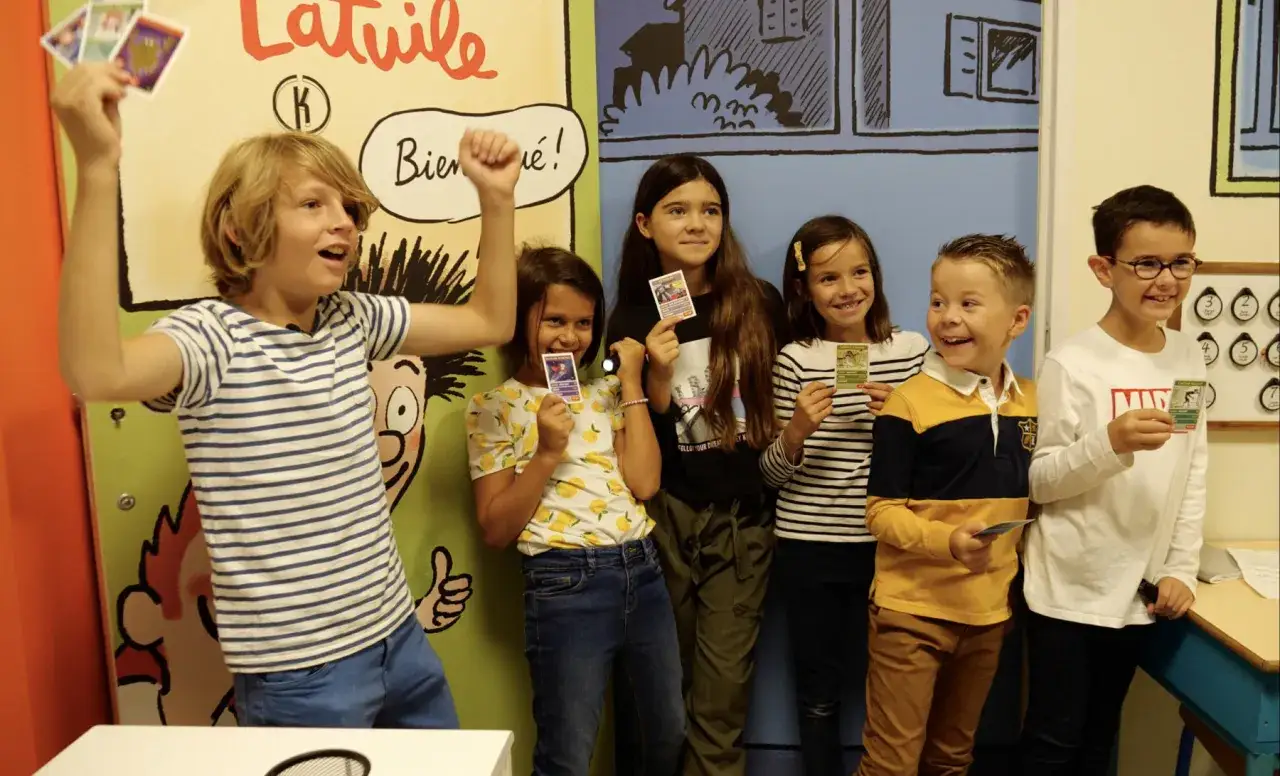 Des enfants heureux participent à un escape game, tenant des cartes colorées. L'un d'eux lève les bras en signe de victoire.