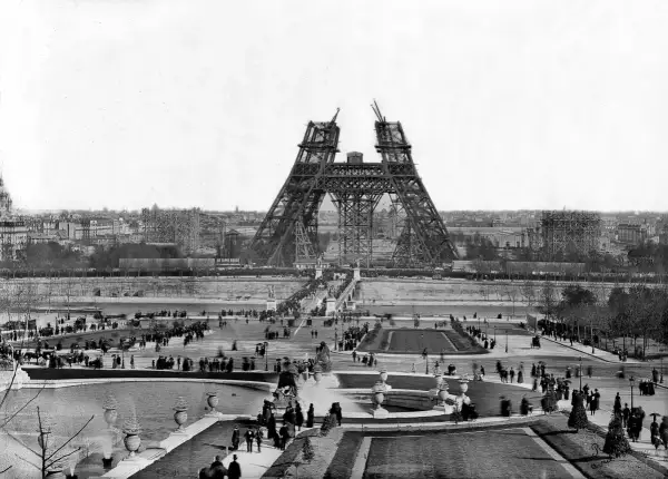 La Tour Eiffel en construction, montrant les matériaux métalliques assemblés. Des foules observent l'avancée des travaux sur le Champ de Mars.