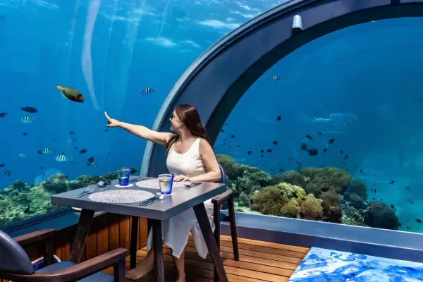 Une femme pointe un poisson dans un restaurant sous la mer, entourée de coraux et de vie marine.