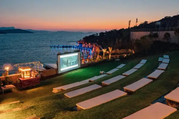 Un magnifique **cinéma plein air** au bord de la mer, avec un écran projetant un film, des sièges sur l'herbe et un chariot de pop-corn.