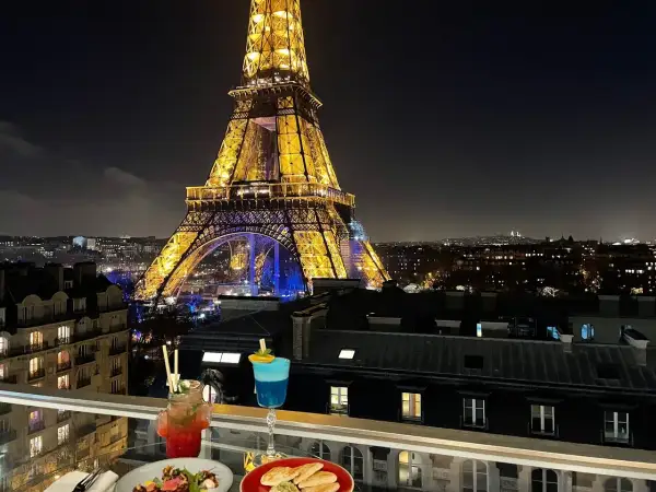 Vue nocturne de Paris depuis un balcon avec des cocktails et des tapas, la Tour Eiffel illuminée en arrière-plan. C'est le 10ème ciel !