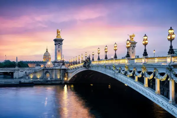 Le **pont Alexandre III** s'illumine sous un ciel crépusculaire aux teintes roses et violettes, reflétant sa splendeur dorée sur la Seine.