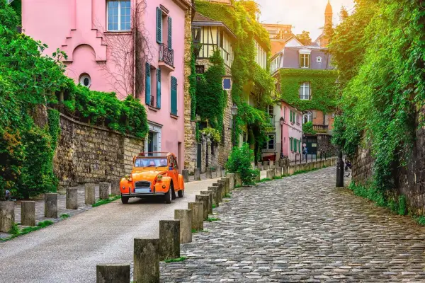 Une 2CV orange monte une rue pavée bordée de maisons roses et vertes. Parfait pour savoir que faire ce dimanche.