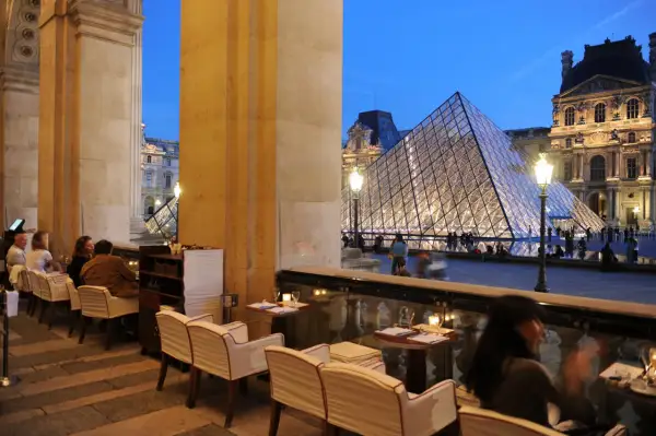 Terrasse du Café Marly, vue sur la pyramide du Louvre illuminée, avec des clients attablés.
